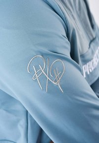 Ljust blått tyg med en slät textur, prytt med en silverbroderad logotyp på ärmen, som visar stiliserade initialer och kurvor.