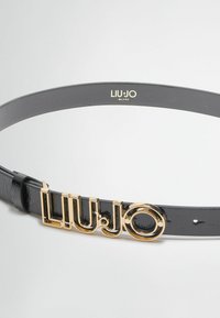Ceinture en cuir noir avec une finition brillante, une boucle logo en ton or proéminente et un design lisse et épuré. Réglable avec des trous.