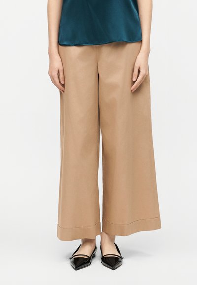 Femme portant un pantalon large beige, un haut en satin teal et des ballerines noires à bout pointu, debout devant un fond clair uni.