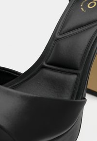 Musta korkokenkä, jossa on sileä nahkainen remmi ja pehmustettu pohjallinen. Muotoilussa on lisäksi beige sisävuori ja logo.