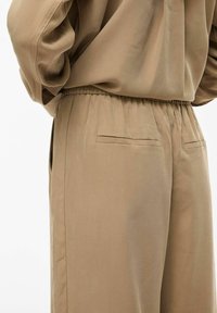 Pantalon beige ample avec ceinture élastique et poches passepoilées à l'arrière, porté avec un haut beige assorti rentré dedans.