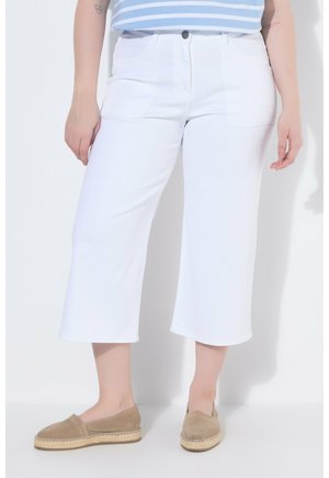 Kvinde iført hvide cropped bukser med vide ben, blå- og hvidstribet skjorte og beige slip-on sko, stående mod en ensfarvet baggrund.