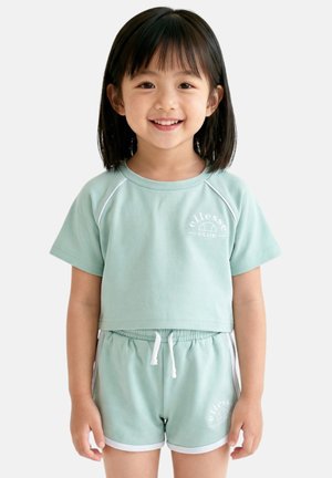 Jeune fille souriante, portant un haut vert menthe à manches courtes et un short assorti avec une bordure blanche et un cordon, debout sur un fond blanc.