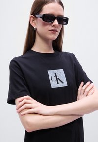 Μαύρο βαμβακερό μπλουζάκι με τετράγωνο γκρι λογότυπο που απεικονίζει το "ck." Το μοντέλο φοράει μαύρα ορθογώνια γυαλιά ηλίου. Ομαλή υφή, κοντά μανίκια.