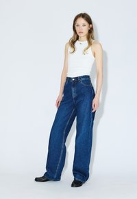 Jeans in denim blu a vita alta e gamba larga con cuciture a contrasto, tasche laterali e chiusura con bottone; indossati con un top bianco aderente senza maniche.