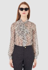 Blusa trasparente con motivo leopardato e un frontale arricciato, abbinata a pantaloni scuri a righe sottili. Gli accessori includono occhiali da sole neri a forma di gatto.