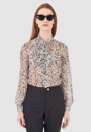 Blusa trasparente con motivo leopardato e un frontale arricciato, abbinata a pantaloni scuri a righe sottili. Gli accessori includono occhiali da sole neri a forma di gatto.