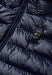 Giubbotto puffy blu navy con una texture liscia e lucida; design trapuntato con sezioni cucite; etichetta attaccata con testo giallo a contrasto.