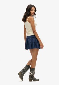 Top sin mangas beige, falda de capas azul marino y botas de vaquero negras con estrellas plateadas y tachuelas. La falda tiene una cintura fruncida.