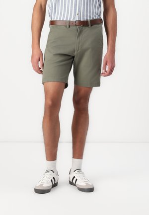 Olivgrüne Shorts mit geradem Schnitt und flachem Bund, kombiniert mit einem braunen Gürtel. Das Modell trägt weiße Sneaker mit schwarzen Streifen und grauen Akzenten.