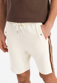 Vita bomullsshorts med dragsko i midjan, med bruna vertikala ränder och silverfärgade knapprapporter på sidofickorna.