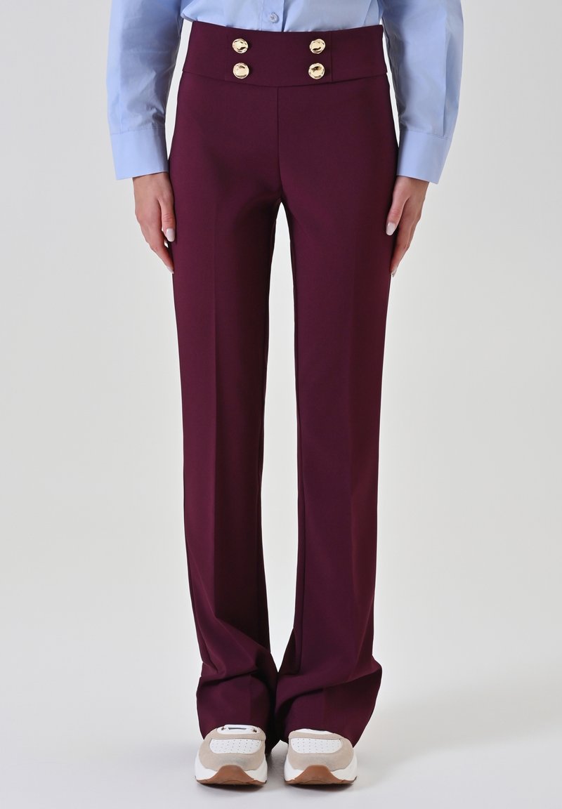 Pantalon sur mesure bordeaux à taille haute, avec des boutons dorés en accent et une texture lisse, associé à des chaussures de sport blanches et grises.