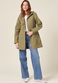 Giacca leggera verde oliva con cappuccio, chiusura con zip e bottoni a pressione. Abbinata a jeans a gamba larga blu chiaro e sneakers bianche.