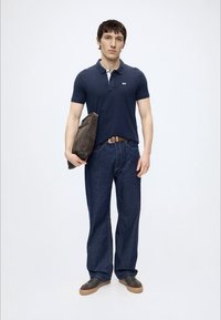 Man standing wearing navy polo shirt, dark blue jeans, brown belt, and sneakers, holding a brown leather bag under his arm.