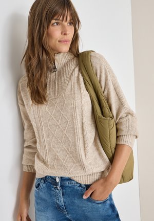Beige Kabelstrickpullover mit hohem Kragen und gerippten Bündchen, kombiniert mit blauen Jeans und einer olivgrünen Stepp-Schultertasche.