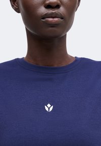 Gros plan sur une personne portant une chemise bleue avec un petit logo floral blanc centré sur la poitrine sur un fond uni.