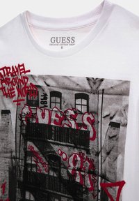 T-shirt bianco Guess con stampa di un edificio urbano in bianco e nero, sovrapposta da testo in stile graffiti rosso che recita "Viaggia per il Mondo" e "Guess."