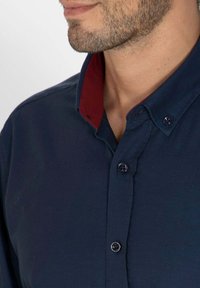 Chemise à boutons bleu marine avec un col intérieur bourgogne contrastant. Texture lisse avec col classique et cinq boutons.