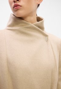 Abrigo de lana beige con un cuello alto, que cuenta con un cierre delantero diagonal y un pin de metal plateado en el lado izquierdo. Textura suave.