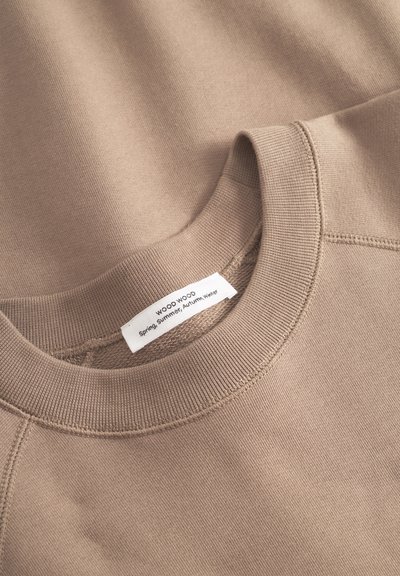 Felpa beige con collo a girocollo a coste, texture morbida e dettagli di cucitura visibili. L'etichetta riporta "WOOD WOOD, Primavera, Estate, Autunno, Inverno."
