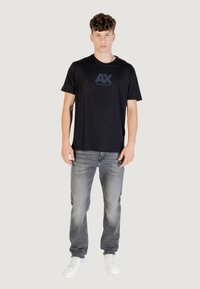 T-shirt noir à manches courtes avec un logo "AX" texturé, associé à un jean gris clair et des baskets blanches, offrant une coupe classique.