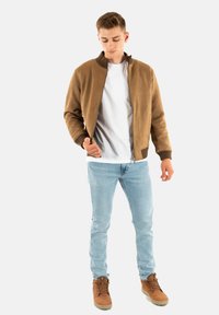 Giubbotto bomber marrone con colletto a costine, abbinato a una T-shirt bianca e jeans azzurri chiari. Stivali marroni con lacci completano l'outfit.