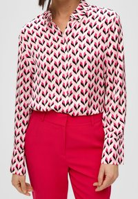 Långärmad blus i rosa med ett fjädermönster i svart och magenta, tillverkad av ett glatt tyg. Tillsammans med högmidjade rosa byxor.