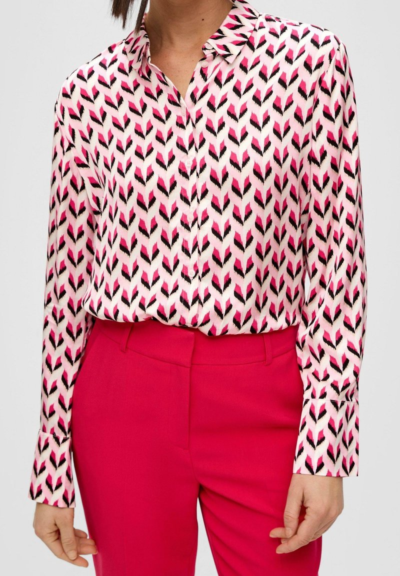 Långärmad blus i rosa med ett fjädermönster i svart och magenta, tillverkad av ett glatt tyg. Tillsammans med högmidjade rosa byxor.