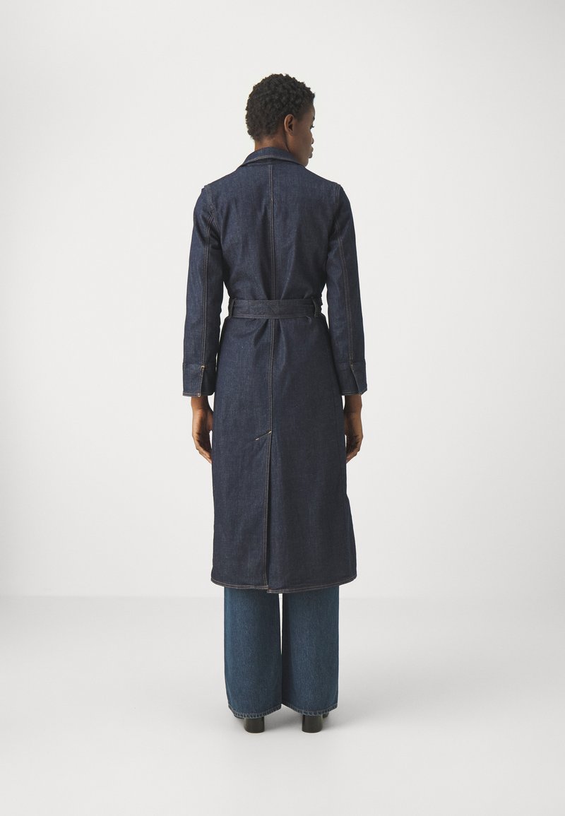 Cappotto di denim blu scuro con maniche lunghe, vita con cintura e un'apertura sul retro. Indossato sopra jeans blu a gamba larga. Texture liscia.