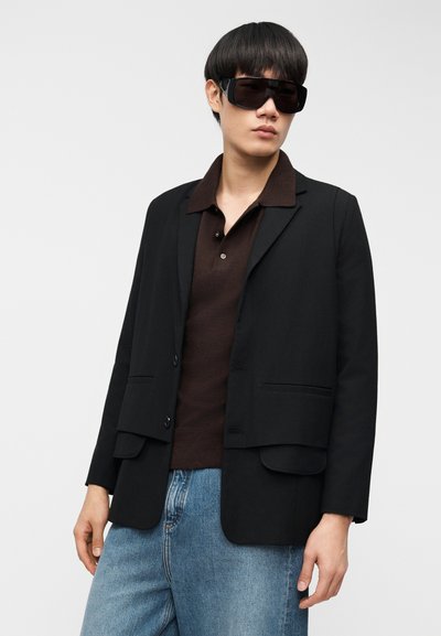 Μαύρο σακάκι με δύο μπροστινές τσέπες, φορεμένο πάνω από σκούρο καφέ πόλο πουκάμισο, συνδυασμένο με ανοιχτό μπλε τζιν. Το μοντέλο φοράει μαύρα, υπερμεγέθη γυαλιά ηλίου.