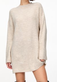 Robe-pull en maille beige avec une coupe ample, manches longues, encolure ronde et un ourlet qui remonte sur un côté. Texture douce.