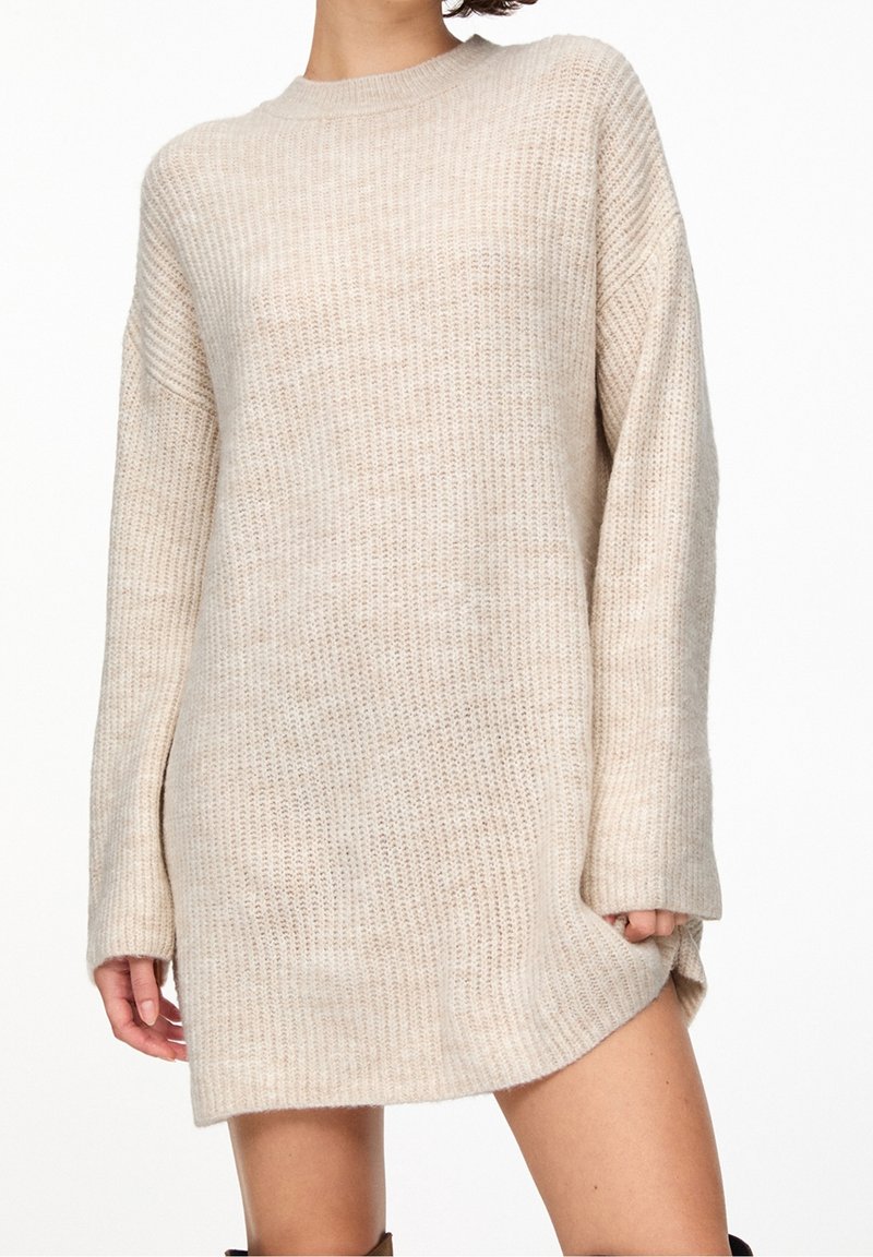 Robe-pull en maille beige avec une coupe ample, manches longues, encolure ronde et un ourlet qui remonte sur un côté. Texture douce.