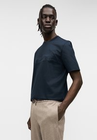 Σκούρο μπλε κοντομάνικο t-shirt με ανάγλυφο σχέδιο αυτοκινήτου, κατασκευασμένο από λείο ύφασμα. Συνδυασμένο με μπεζ παντελόνι με κορδόνι στη μέση και πλαϊνές τσέπες.