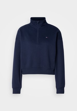 Sweatshirt cropped navy avec un col haut et une fermeture éclair quart. Présente des poignets et un ourlet côtelés, avec un petit logo sur la poitrine. Texture douce.