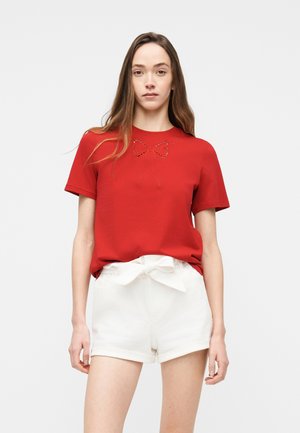 Femme portant une chemise rouge à manches courtes avec un petit nœud comme motif et un short blanc taille haute noué avec une ceinture en tissu, debout devant un fond uni.