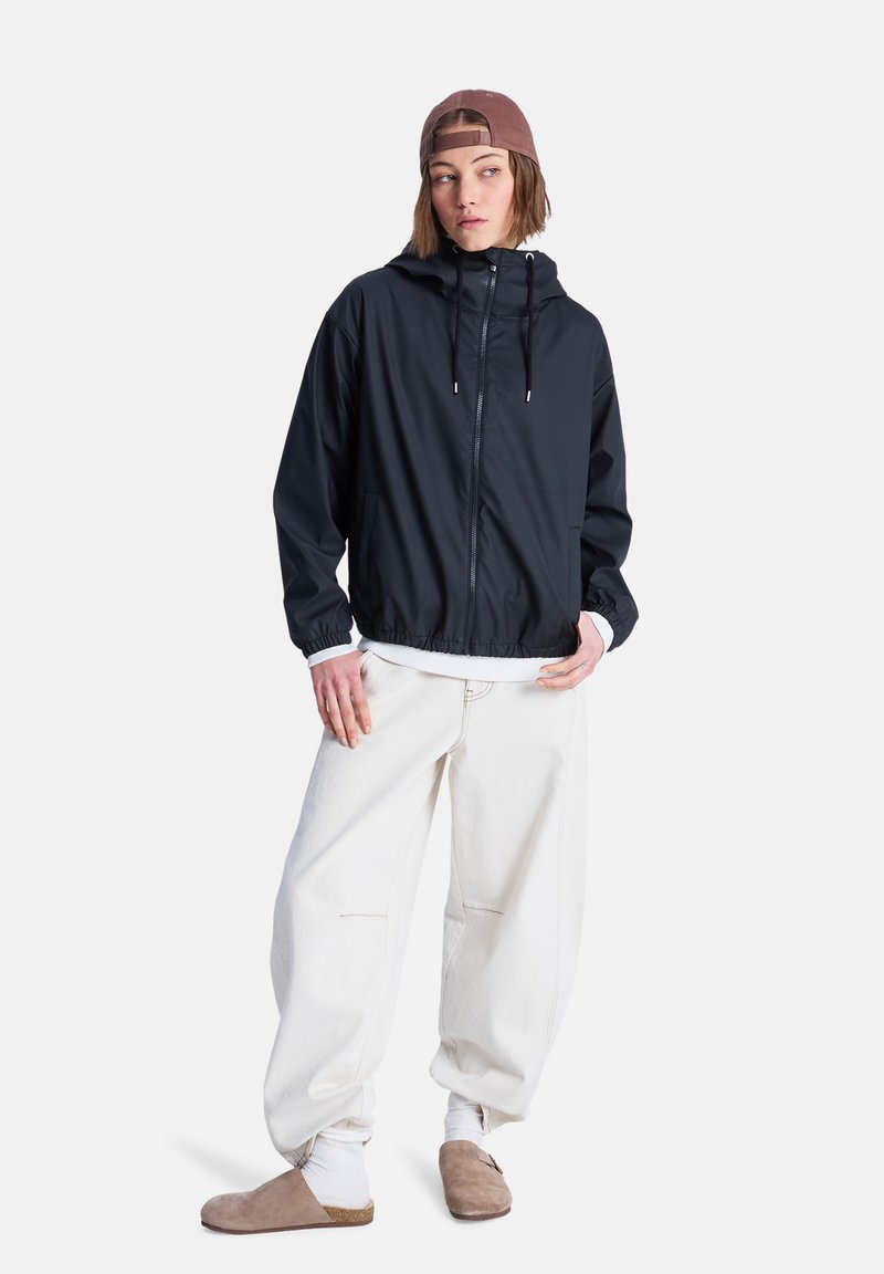 Μαύρο μπουφάν με κουκούλα και φερμουάρ μπροστά, ελαστικά μανίκια και πλαϊνές τσέπες, συνδυασμένο με φαρδιά λευκά παντελόνια και μπεζ παπούτσια τύπου slip-on.