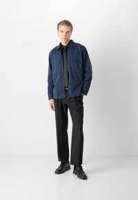 Merevaigist sinine jakk kahe plaastriga taskuga, koos mustade kitsaste pükste ja mustade nööbiga kingadega. Mudel seisab ühevärvilise heleda tausta ees.