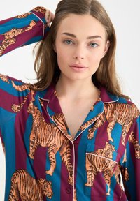 Lange mouw pyjama-top in blauw en bordeauxrood met een overal tijgerprint, voorzien van contrasterende witte piping en een zak aan de linkerkant van de borst.