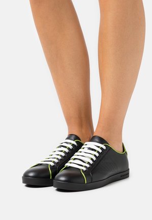 Baskets en cuir noir avec des lacets blancs, présentant des accents verts et un design à bout rond. Les panneaux latéraux texturés ajoutent du détail.