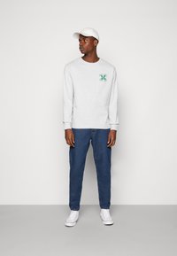 Sweatshirt cinza de mangas longas com logotipo verde, combinado com jeans de denim azuis e ténis brancos. O boné também é branco e sem adornos.