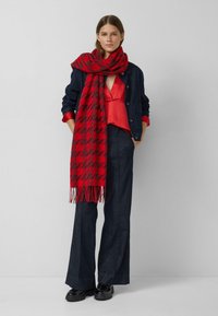 Frau trägt eine rote Bluse, eine dunkle Jeansjacke und Hose, schwarze Schuhe sowie einen großen roten Schal mit Muster und steht mit den Händen in den Taschen.