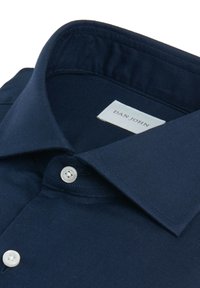Camisa azul oscuro con cuello y botones blancos, doblada cuidadosamente, mostrando la etiqueta interior con el nombre de la marca "Dan John."