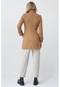 Abrigo de lana marrón con un diseño ajustado, costuras verticales y puños abotonados. Combinado con pantalones gris claro y botas negras de tobillo.