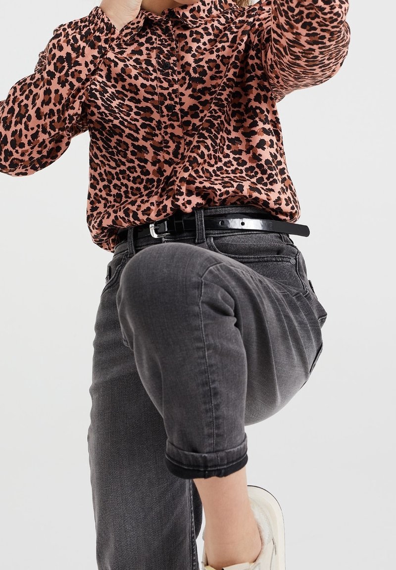 Personne portant un chemisier rose et noir à motif léopard, un jean gris foncé retroussé, une ceinture noire et des baskets blanches avec un genou levé.
