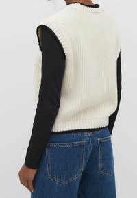 Veste en tricot épais de couleur blanc cassé, avec des bords côtelés et une bordure noire, portée sur un haut noir à manches longues ; associée à un jean en denim bleu.