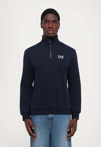 Mørkeblå halv-zip sportstrøye laget av mykt stoff, med høy krage, lange ermer, ribbede mansjetter, og en hvit logo på brystet.
