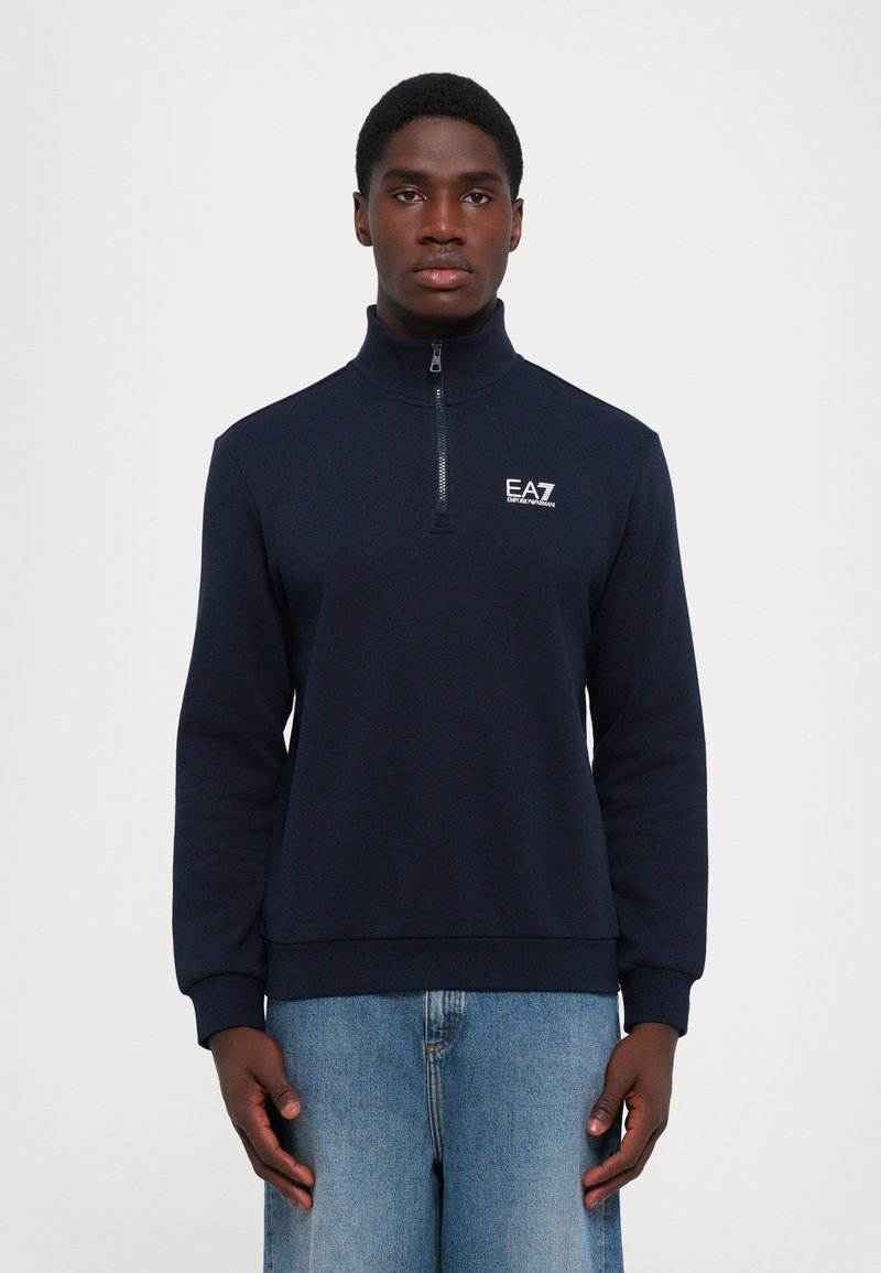 Mørkeblå halv-zip sportstrøye laget av mykt stoff, med høy krage, lange ermer, ribbede mansjetter, og en hvit logo på brystet.