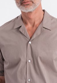 Košile s krátkým rukávem světle hnědé barvy, zapínaná na knoflíky, vyrobená z hladké látky, s klasickým límcem a bílými knoflíky na přední straně.