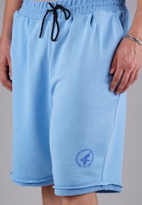 Light blue cotton shorts with an elastic waistband and black drawstring. Features a circular logo in navy on the left leg. Soft texture.