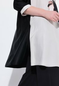 Zwart-witte tuniek met een hoge zijsplit, gemaakt van gladde, glanzende stof en contrasterende texturen; korte mouwen met witte afwerking.